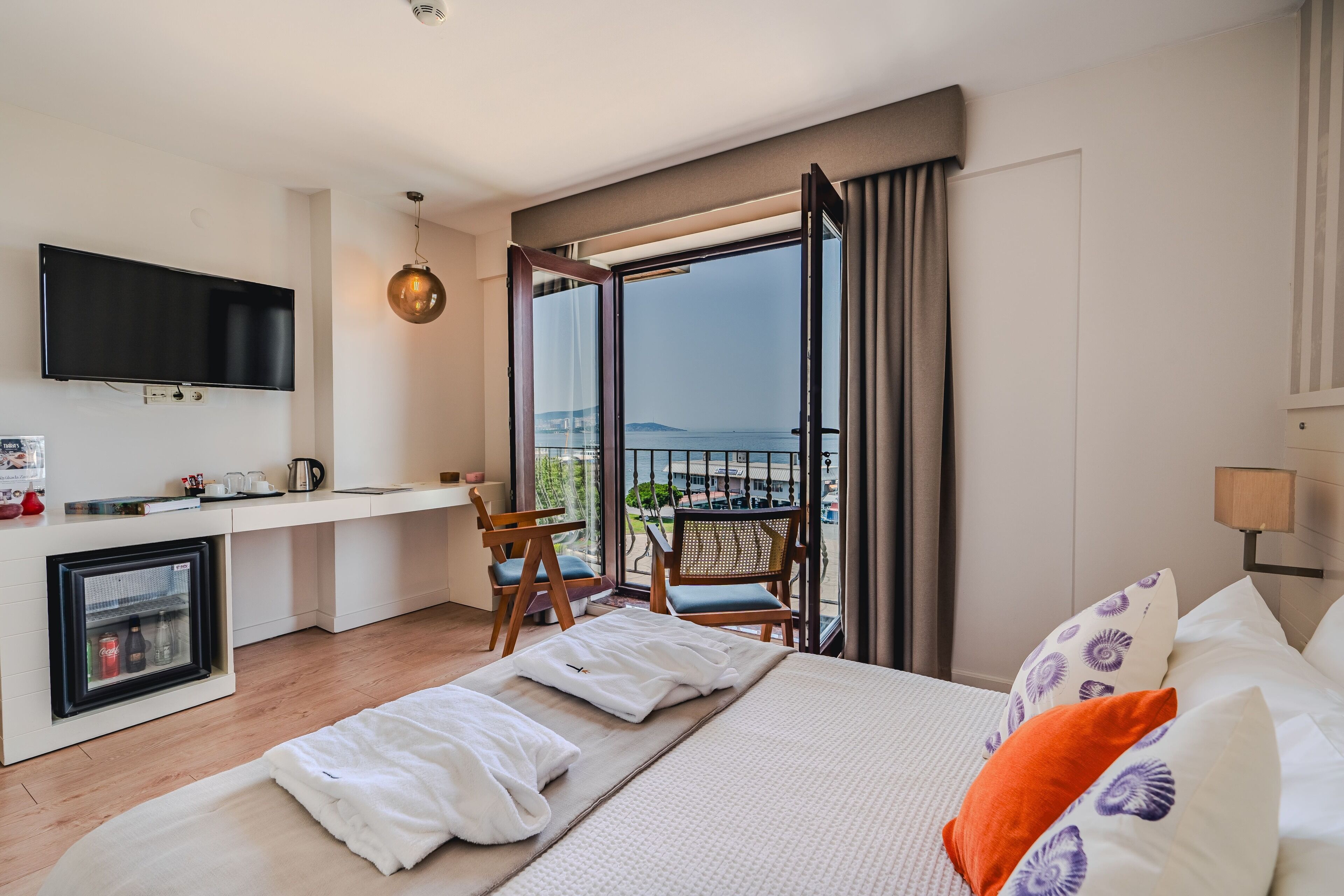A hotel room with a bed, a TV, a small kitchenette, a balcony with a view of the sea, and a chair.