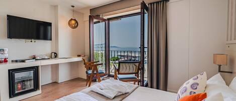 A hotel room with a bed, a TV, a small kitchenette, a balcony with a view of the sea, and a chair.