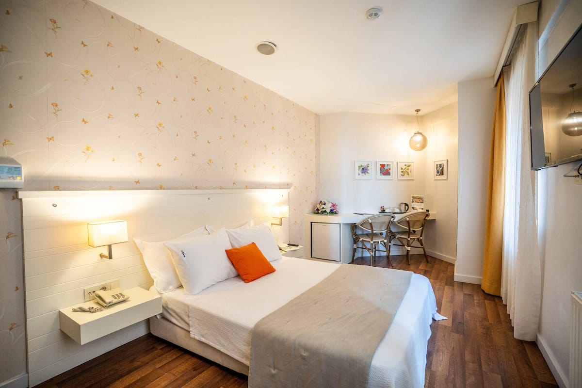 A hotel room with a bed, bedside table with a phone, a desk with a chair, and a small kitchenette.