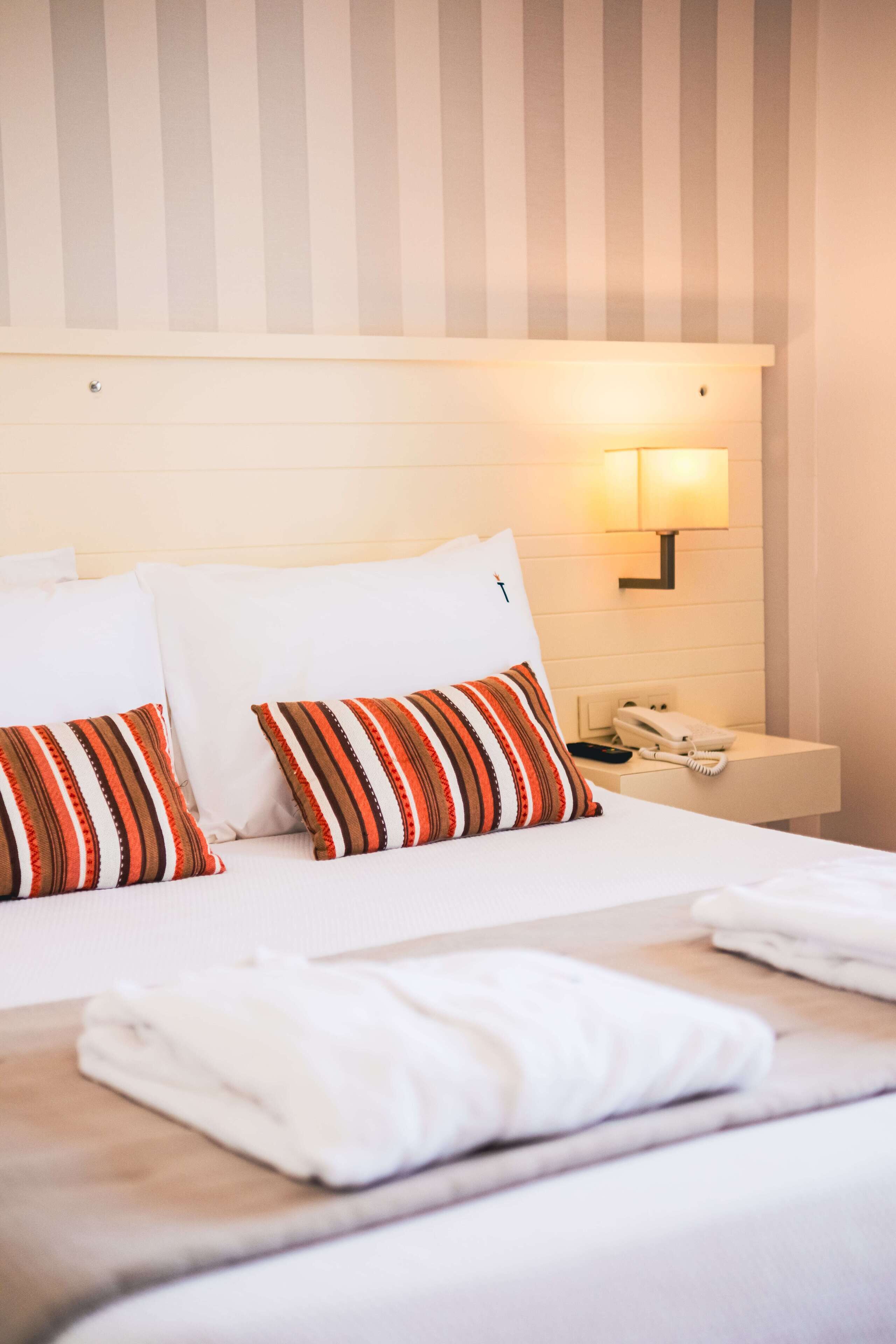 A hotel room with a bed, two striped pillows, a bedside table with a phone, and a wall-mounted lamp.