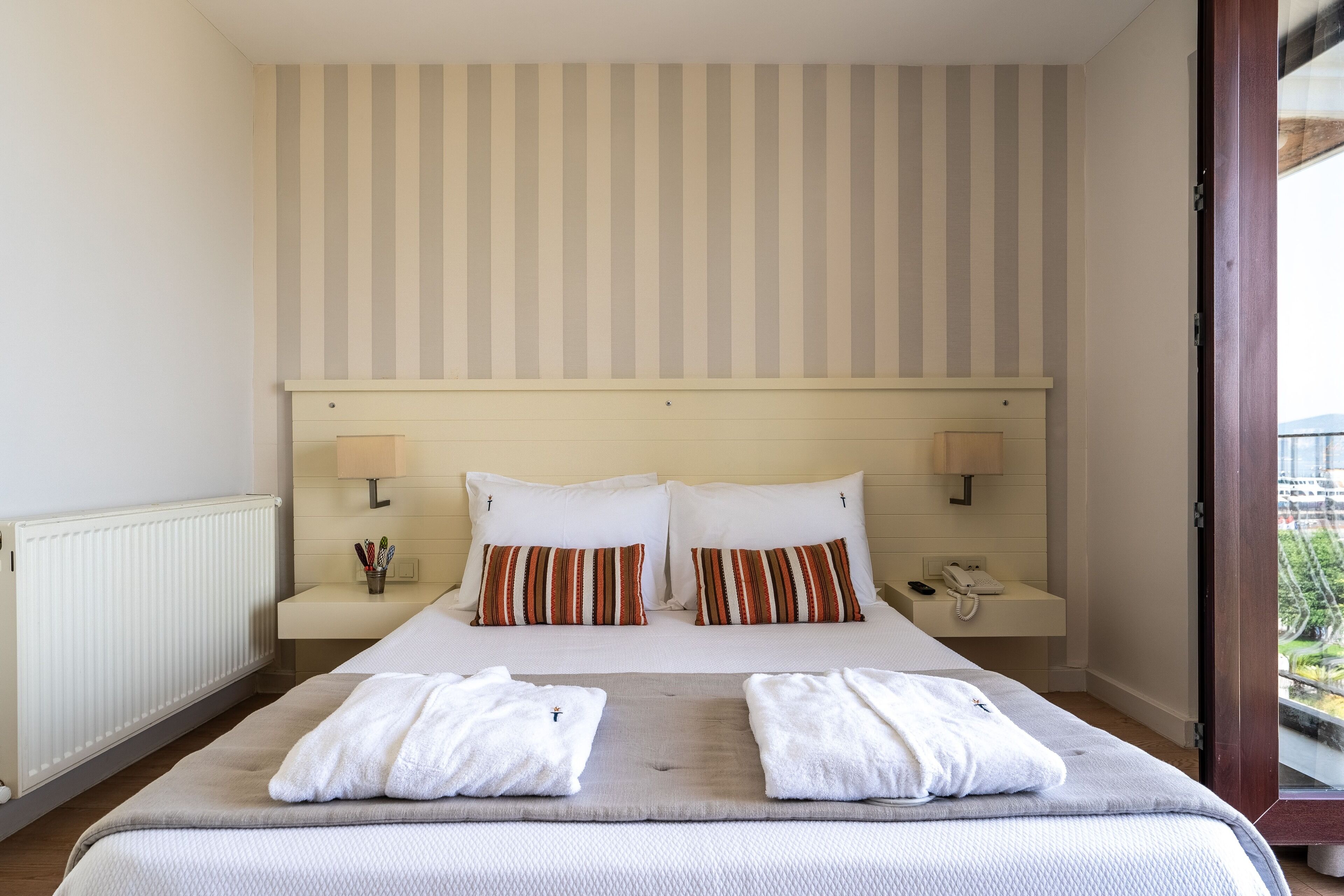 Habitación de hotel con una cama grande, cabecera a rayas, lámparas de noche y vistas al exterior.
