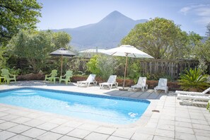 A swimming pool with lounge chairs and umbrellas, surrounded by a wooden fence and greenery, with a mountain in the background. - Andelomi Forest Lodge (Storms River)
