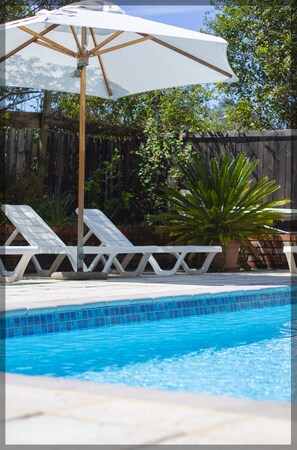 A swimming pool with a wooden deck, white lounge chairs, and a white umbrella.