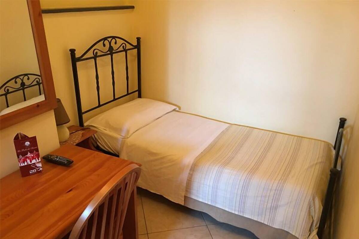 Una habitación pequeña con una cama individual, un escritorio de madera y una silla, una mesita de noche y una lámpara.