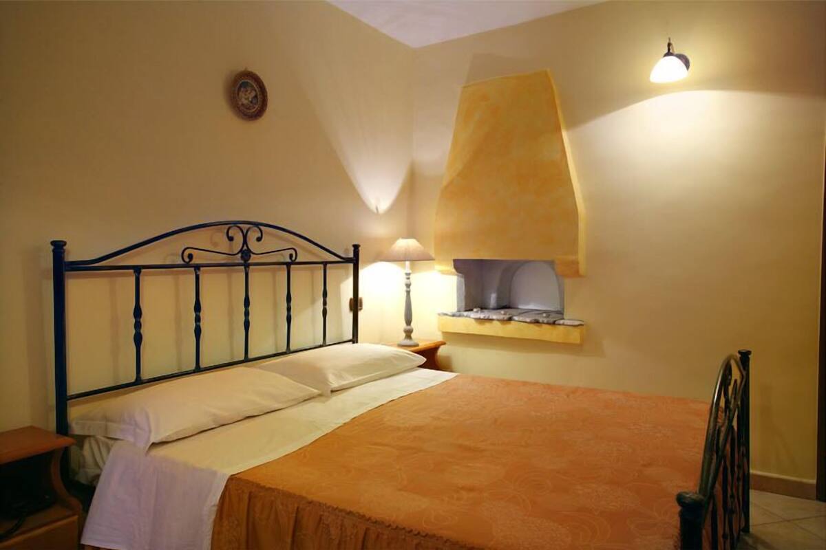 Un dormitorio con cama, mesita de noche, lámpara, estante en la pared y luz de techo.