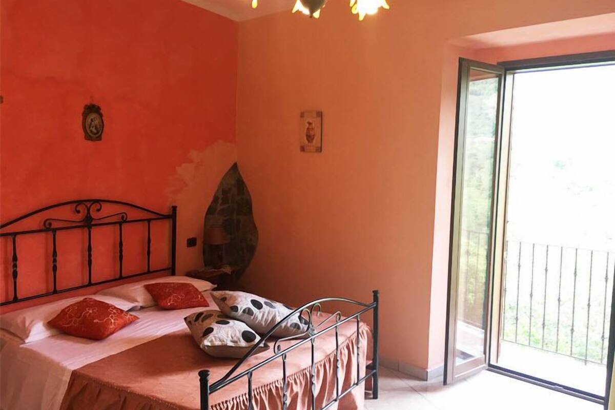 Un dormitorio con cama, almohadas, un reloj de pared y un cuadro. Se ve una ventana con vistas a la vegetación.