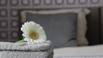 Une fleur blanche posée sur une serviette pliée, dans une pièce dont le papier peint est à motifs et où se trouve un lit avec une tête de lit blanche.