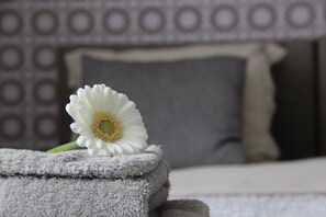 A white flower on a folded towel in a room with patterned wallpaper and a bed with a white headboard. - B&B at Room's (Ypres)