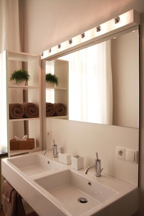 A modern bathroom with a large mirror, two sinks, and a shelf with towels and a plant. - B&B at Room's (Ypres)