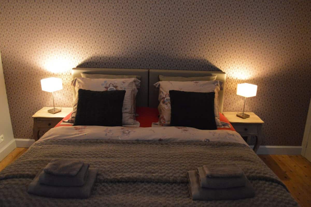 A neatly made bed with two pillows, a blanket, and two folded towels. There are two bedside tables with lamps on either side of the bed.