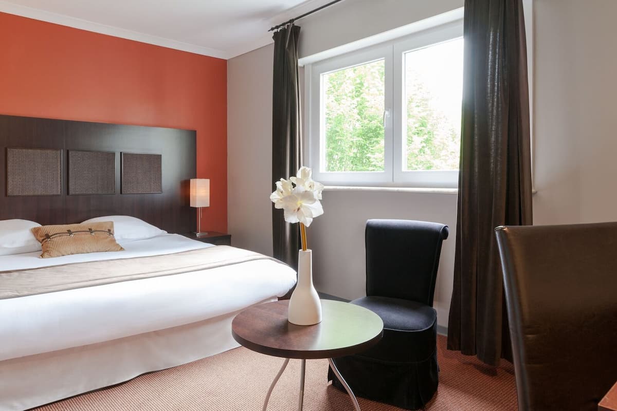 Habitació d'hotel amb llit, una cadira, taula petita i finestra amb cortines.