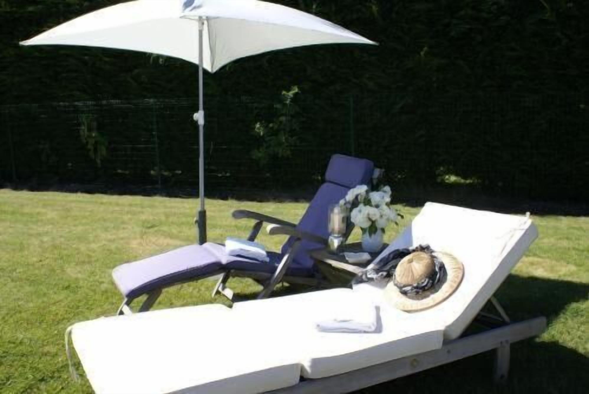 Une chaise longue avec un parasol blanc, un vase rempli de fleurs et un chapeau posé sur la chaise longue.