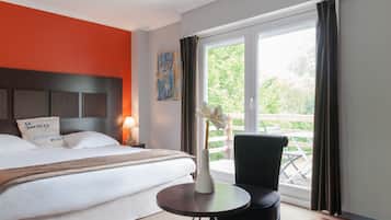 A hotel room with a bed, a round table, a chair, a vase with flowers, a painting, and a view of the outdoors.
