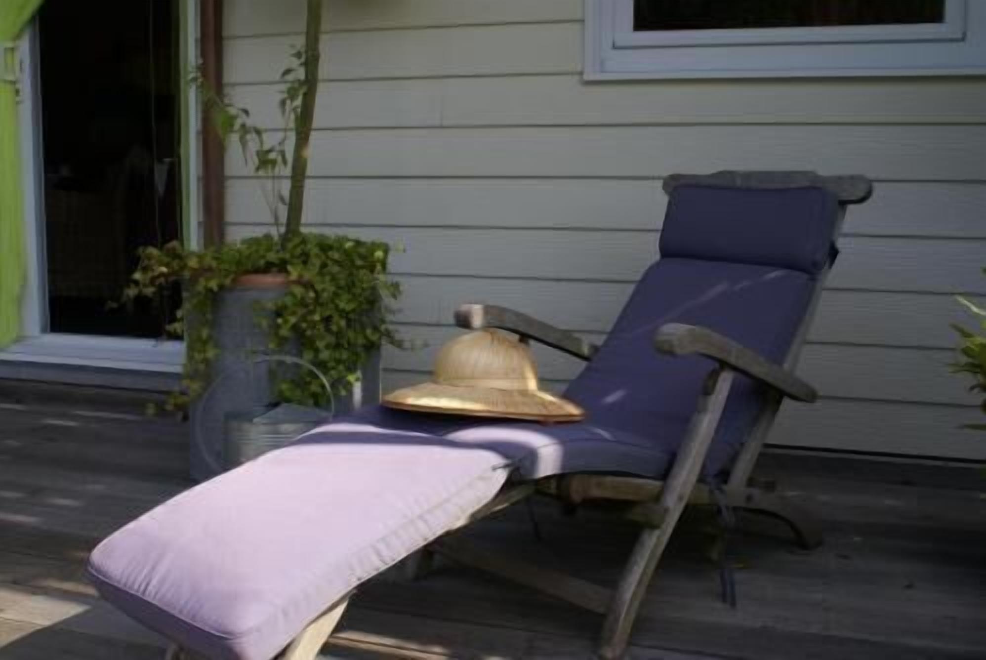 A wooden lounge chair with a cushion, positioned on a porch with a potted plant and a window in the background.