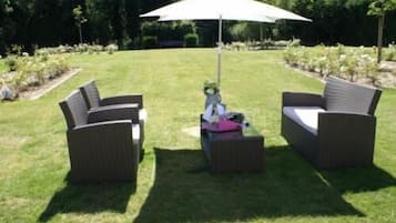 An outdoor patio area with two wicker sofas and a small table, a white umbrella providing shade, and a well-maintained lawn.