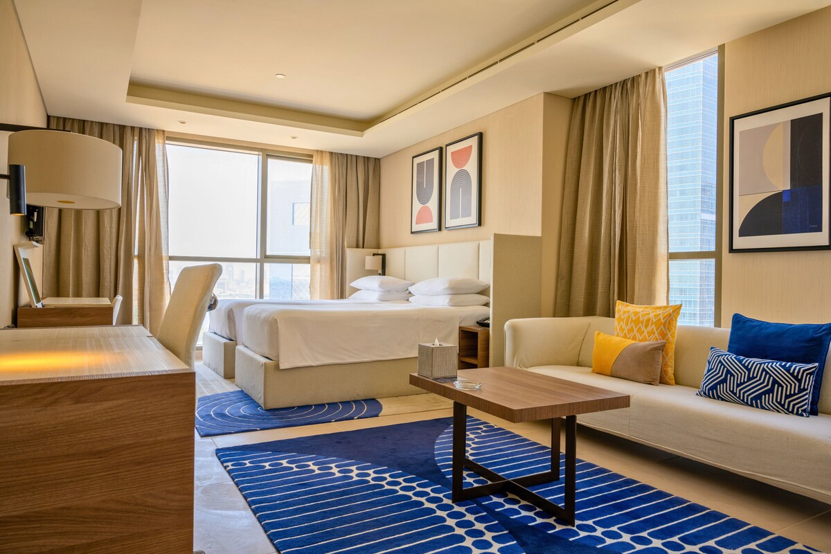 A hotel room with a bed, sofa, desk, and large windows.