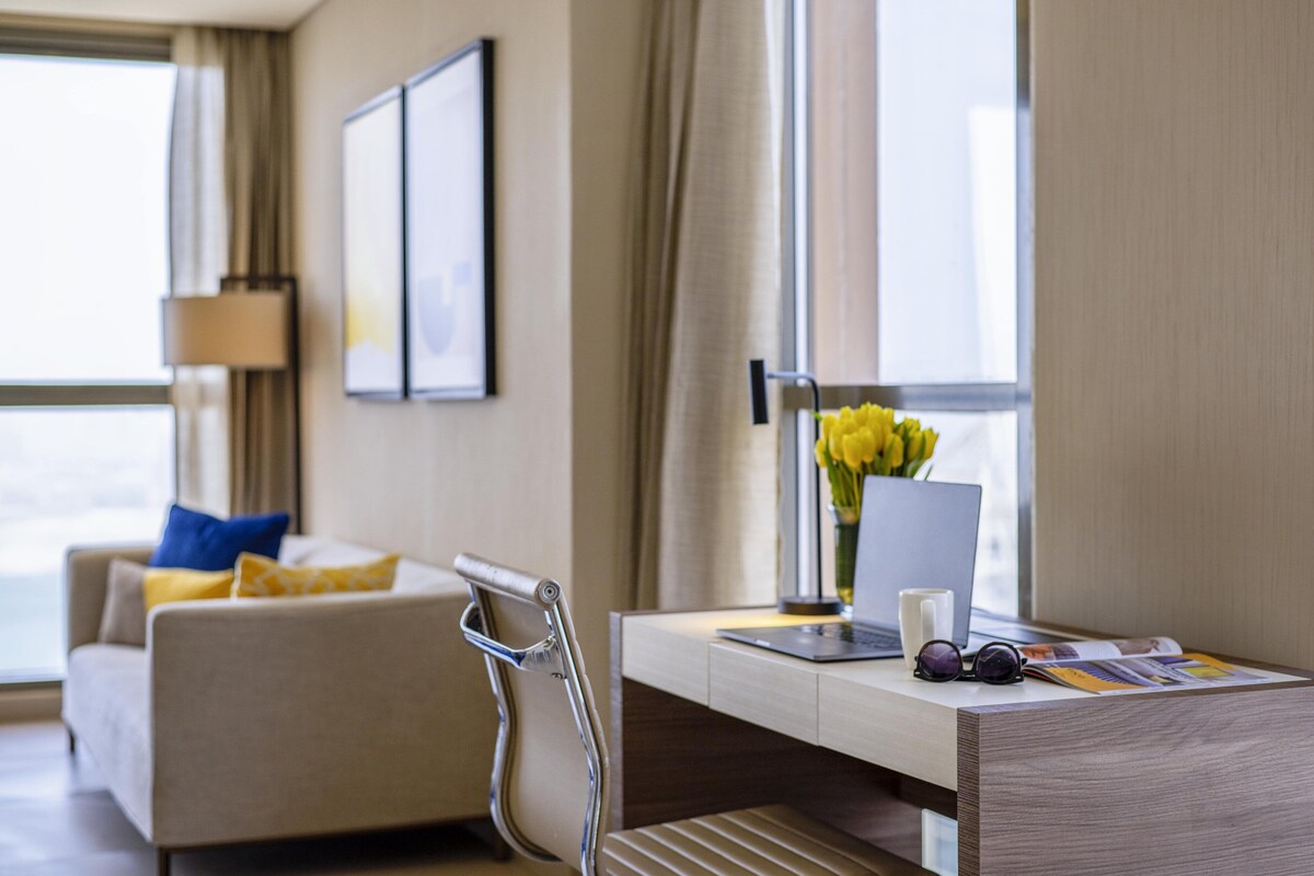 A hotel room with a sofa, a desk with a laptop, a vase with flowers, and a chair.