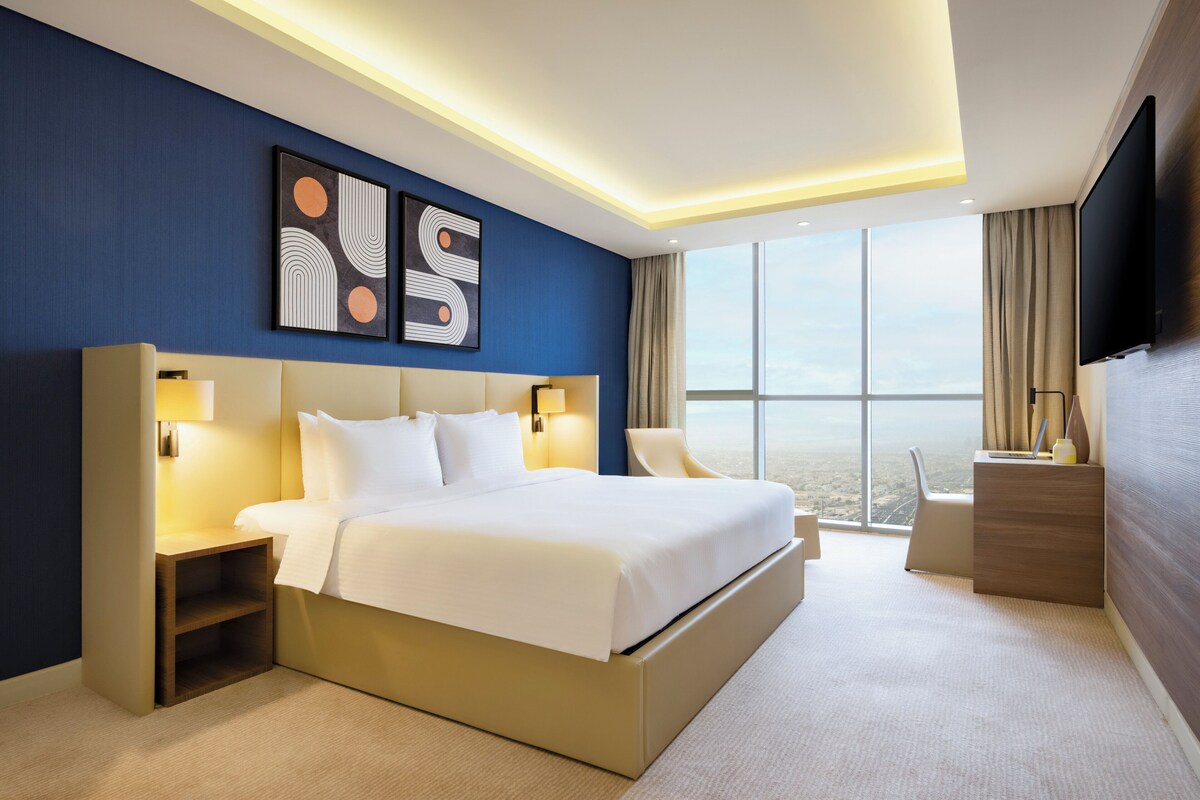 A modern hotel room with a large bed, bedside tables, a flat-screen TV, and a view of the cityscape through large windows.