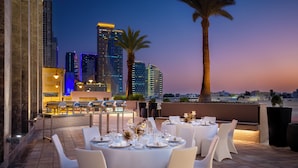 Un espacio al aire libre con mesas dispuestas para una comida, con vistas a la silueta de una ciudad al atardecer.