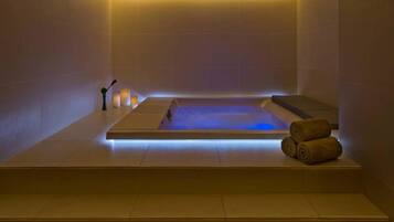A modern spa room with a rectangular hot tub, a small table with candles, and rolled towels on a platform.