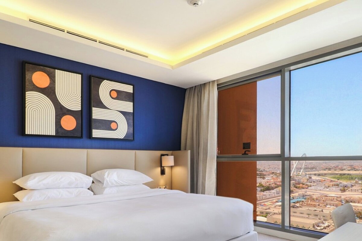 A modern hotel room with a large bed, a desk, and a view of a cityscape through a large window.