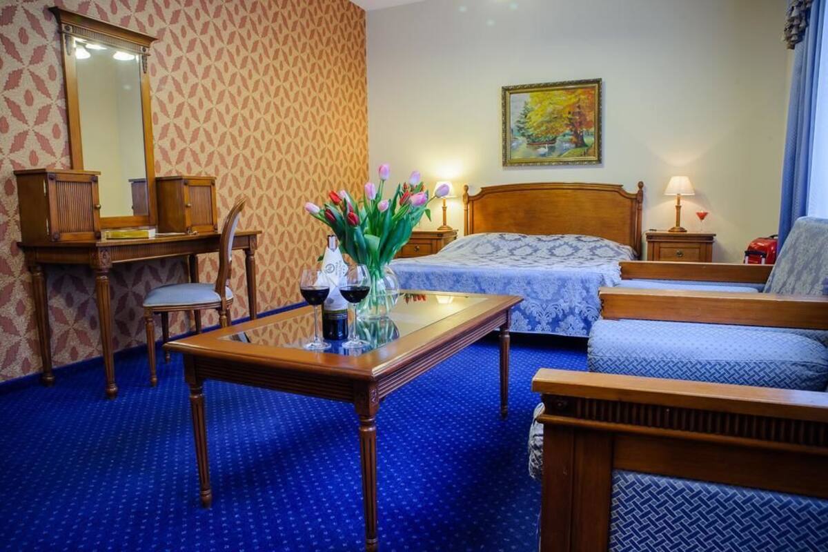 Una habitación de hotel con una cama, un escritorio de madera, una silla, una mesita con un jarrón de flores y un cuadro en la pared.