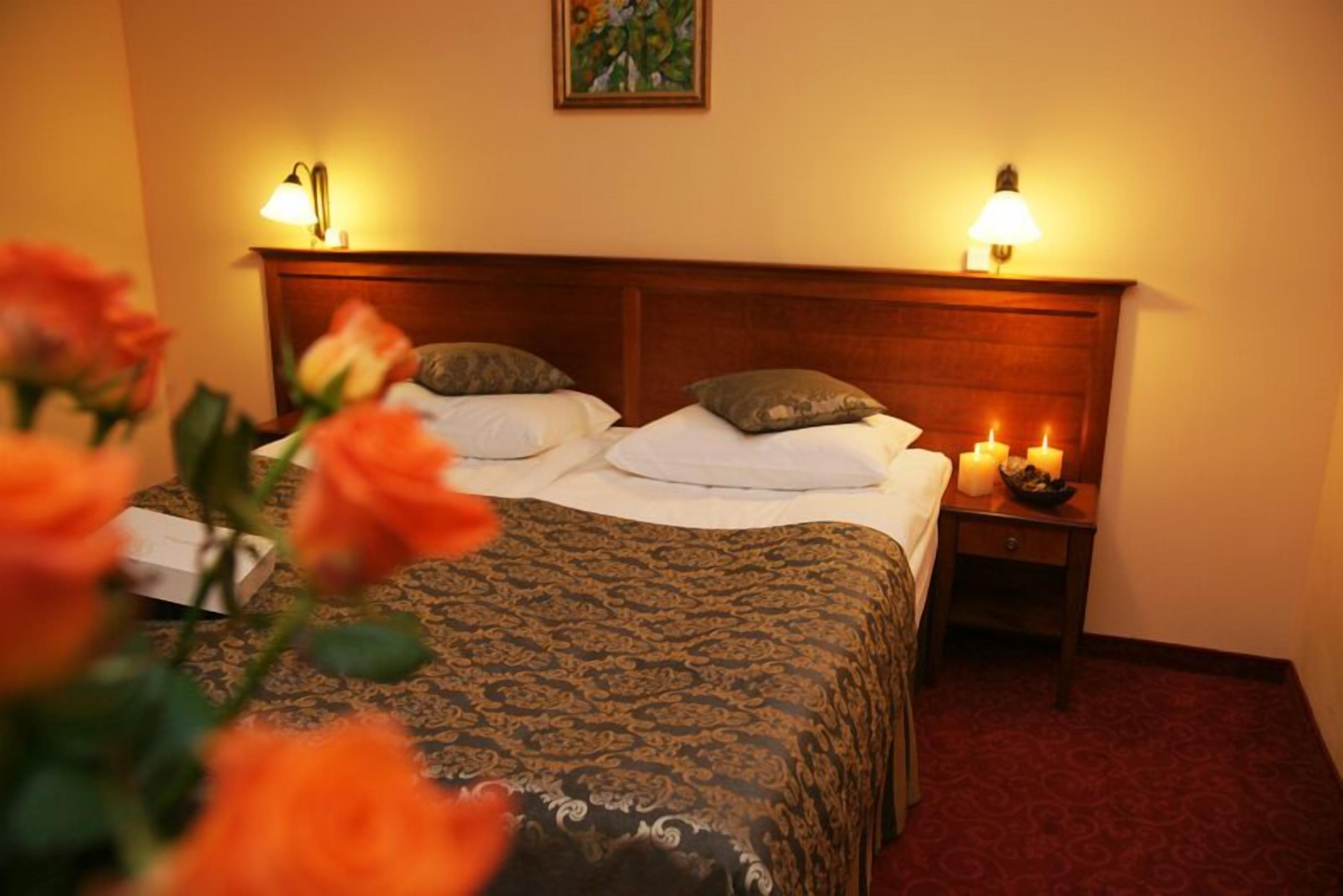 A hotel room with a large bed, two bedside lamps, a nightstand with candles, and a floral arrangement.