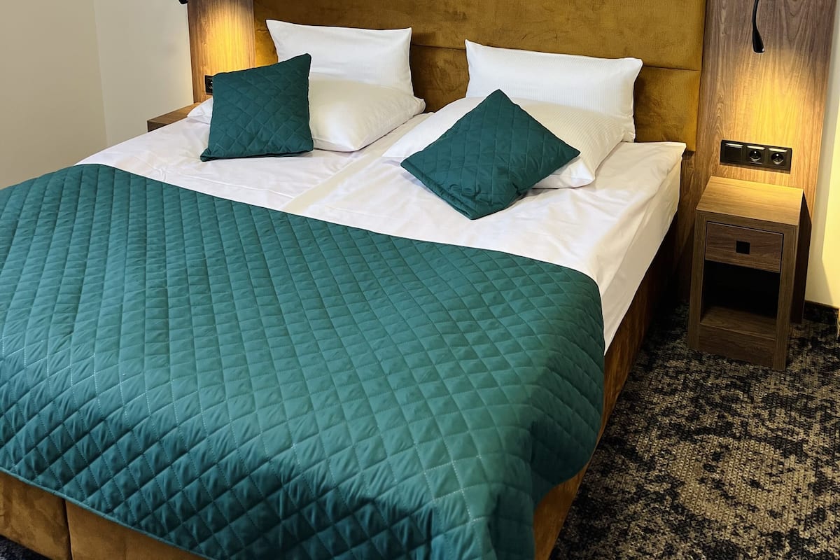 Una cama bien tendida con un edredón verde azulado, dos almohadas blancas y dos almohadas de color verde azulado. Hay dos mesitas de noche con lámparas, y la habitación tiene un techo inclinado.