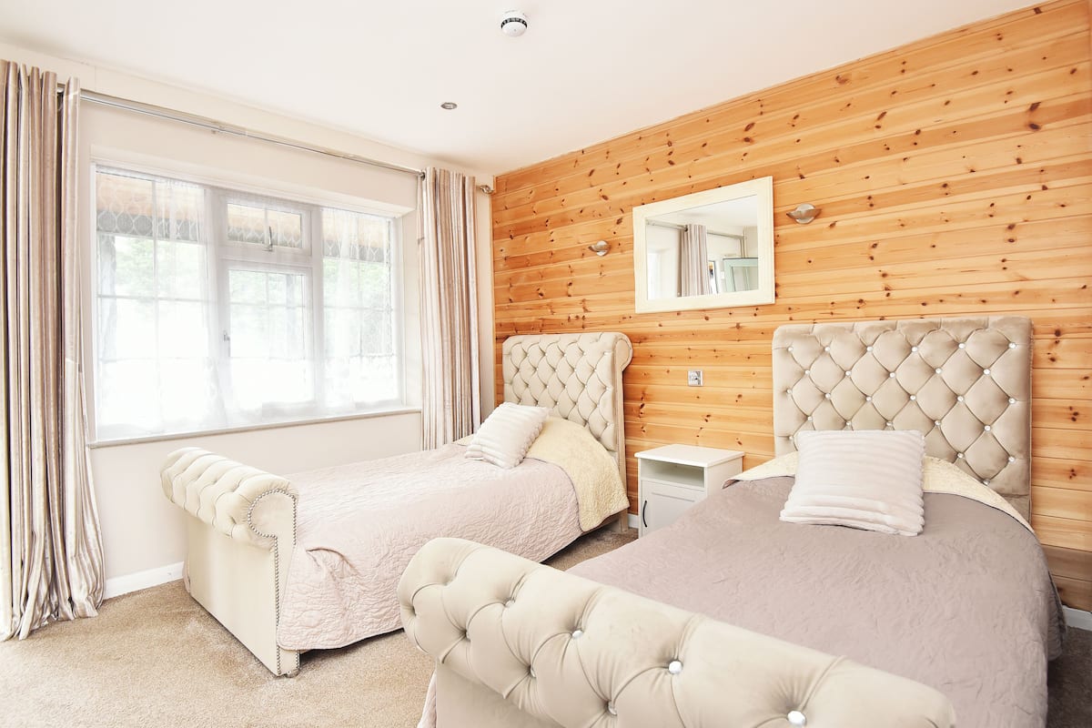Une chambre avec deux lits, des lambris en bois sur les murs, une grande fenêtre avec des rideaux et un miroir au-dessus des lits.