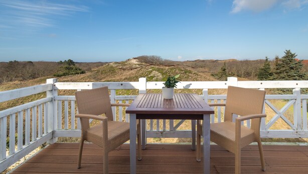 A wooden deck with a white railing, two wicker chairs, and a table with a potted plant.