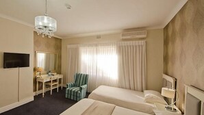 A hotel room with two beds, a desk, a chair, a TV, and a chandelier. - The Russel Hotel (Knysna)