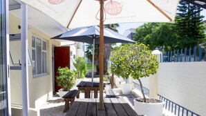 An outdoor patio area with wooden tables and benches, a potted tree, and umbrellas. - The Russel Hotel (Knysna)