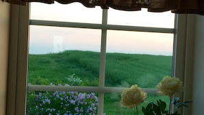 Una ventana con vistas a un campo verde y flores púrpuras, una cesta de mimbre y dos jarrones con flores.