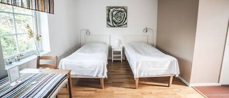 A room with two single beds, a desk with a computer, a window with curtains, and a painting of a rose on the wall.
