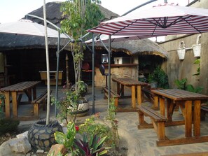 An outdoor patio area with wooden tables and benches, a thatched-roof structure, and a striped umbrella. - Flintstones Backpackers (Lusaka)