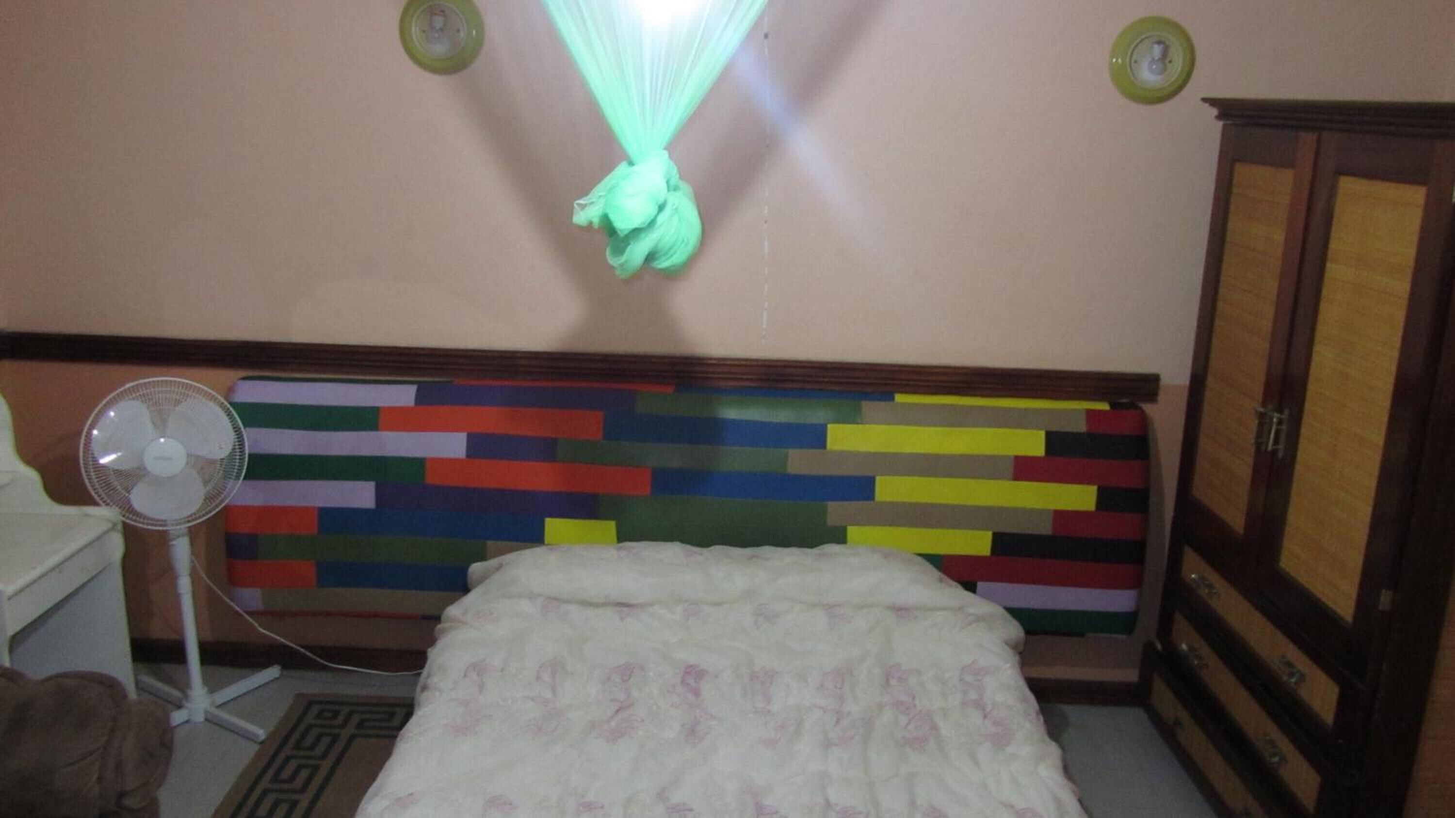 Un dormitorio con un cabecero colorido, una colcha blanca, un ventilador de pie y un armario de madera.