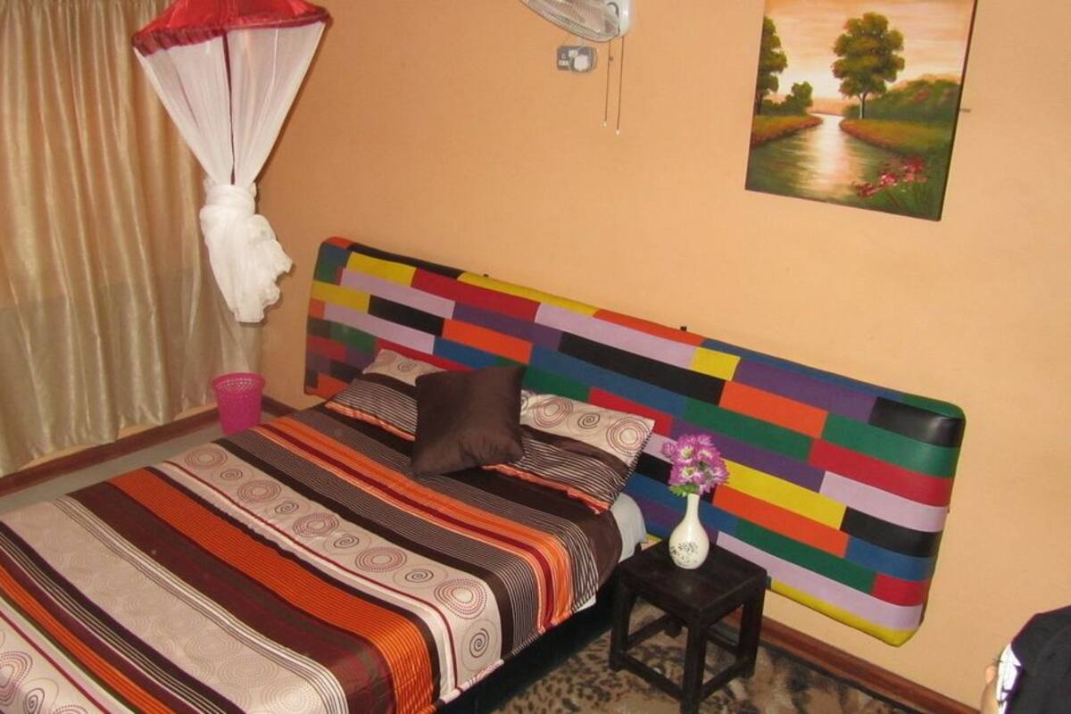 A bed with a multicolored headboard, a bedside table with a vase, and a painting on the wall.