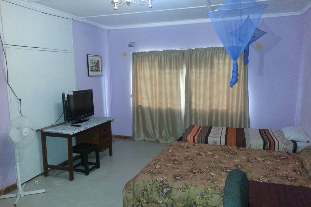 A room with a bed, a desk with a computer, a standing fan, and a window with curtains.