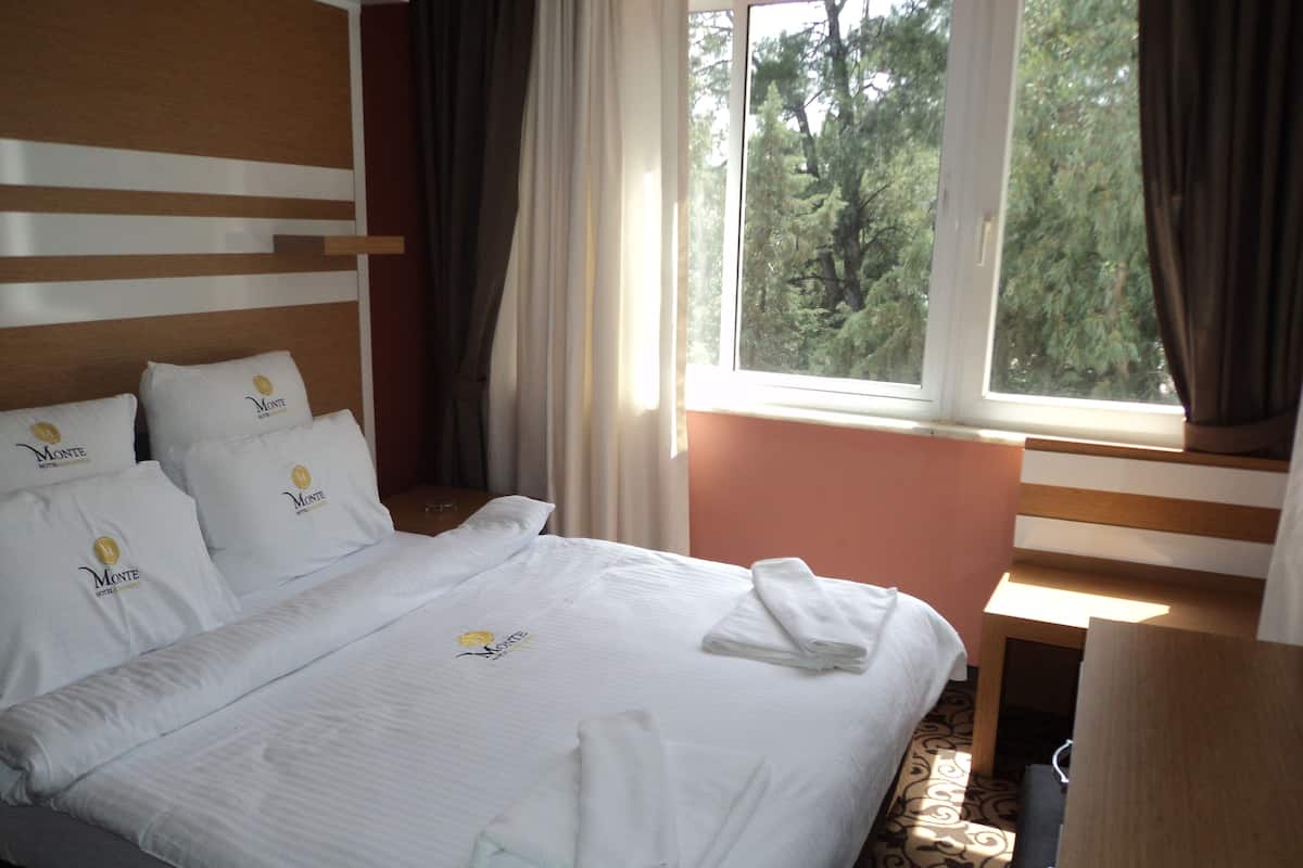 Habitación de hotel con una cama bien hecha, una mesita de noche, una ventana con cortinas y vistas a árboles.