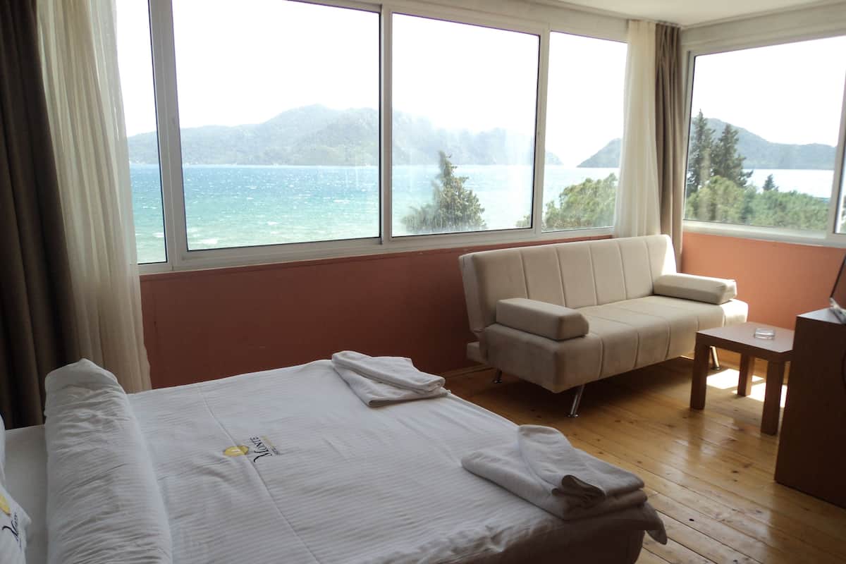 Habitación de hotel con cama, sofá, una mesita y vistas al mar y a las montañas a través de amplios ventanales.
