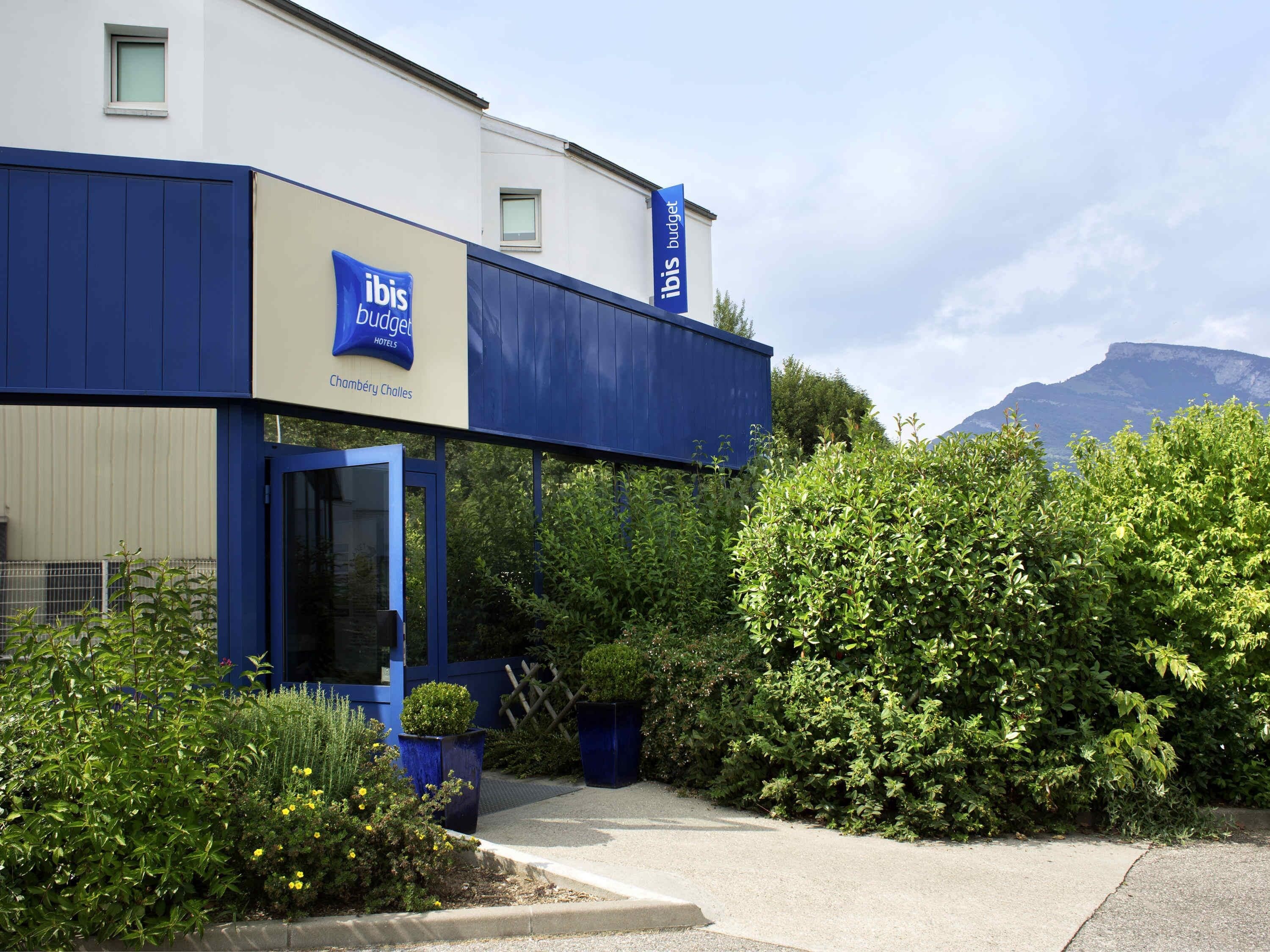 Entrada moderna d'un hotel amb façana blava i blanca, un rètol de 'ibis budget hotels' i una vista d'una muntanya al fons.