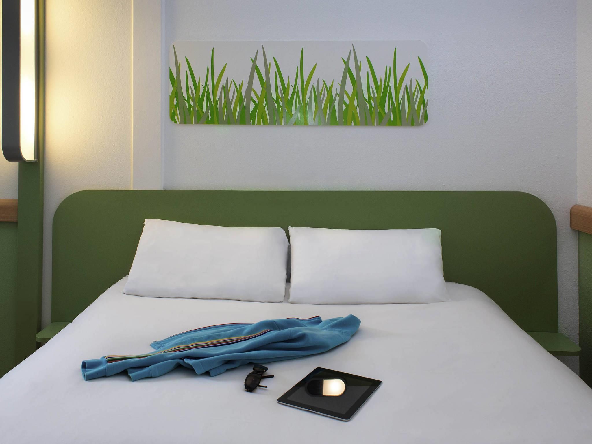 Una cama bien hecha con sábanas blancas y dos almohadas, una chaqueta azul, unas gafas de sol y una tablet sobre la cama.