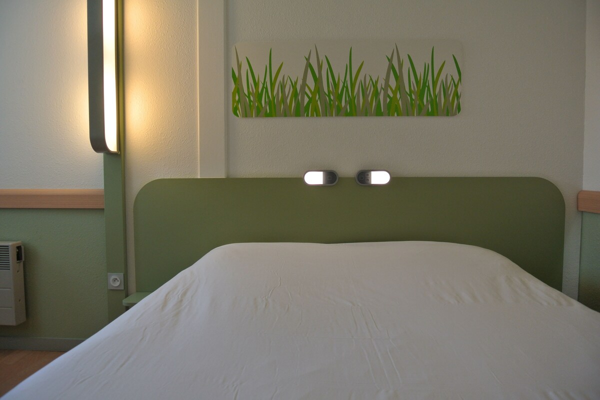 A bed with a green headboard, two bedside lamps, and a wall-mounted grass art piece.