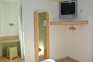 A small, clean room with a wooden stool, a mirror, a television, and a wooden shelf.