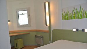 A hotel room with a bed, a small stool, a window, a radiator, and a wall-mounted light fixture. - ibis budget Chambéry Sud Challes les Eaux (Challes-les-Eaux)