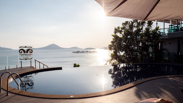 Una piscina infinita con vistas al mar y a las montañas, un cartel que dice "Victoria Cliff" y mobiliario exterior.