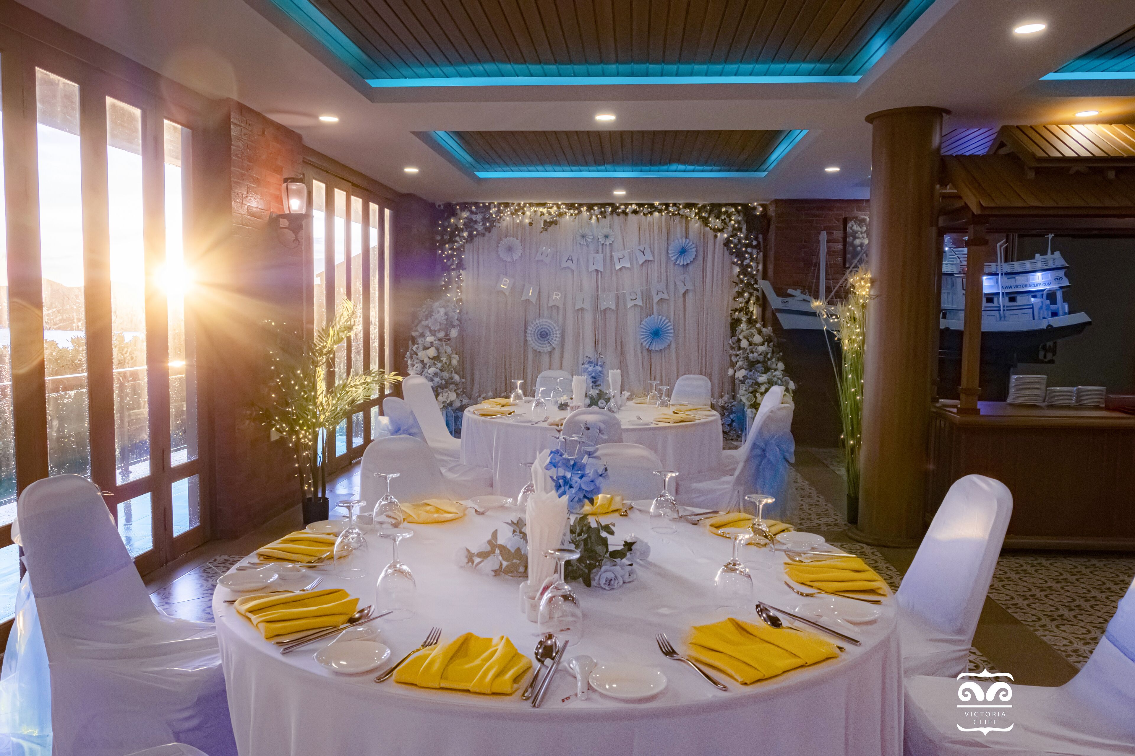 A banquet hall with round tables set for a formal event, featuring white tablecloths, yellow napkins, and floral centerpieces.