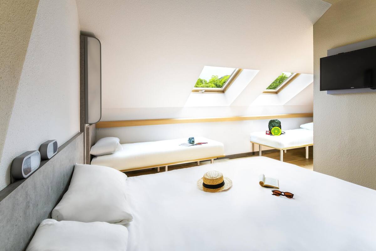 Ein modernes Hotelzimmer mit einem großen Bett, zwei Nachttischen, einem kleinen Schreibtisch und einem großen Fenster mit Blick auf Grünflächen.