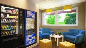 Una habitación con una máquina expendedora, dos sillones azules, una mesita con una planta y una ventana con vistas al verdor.