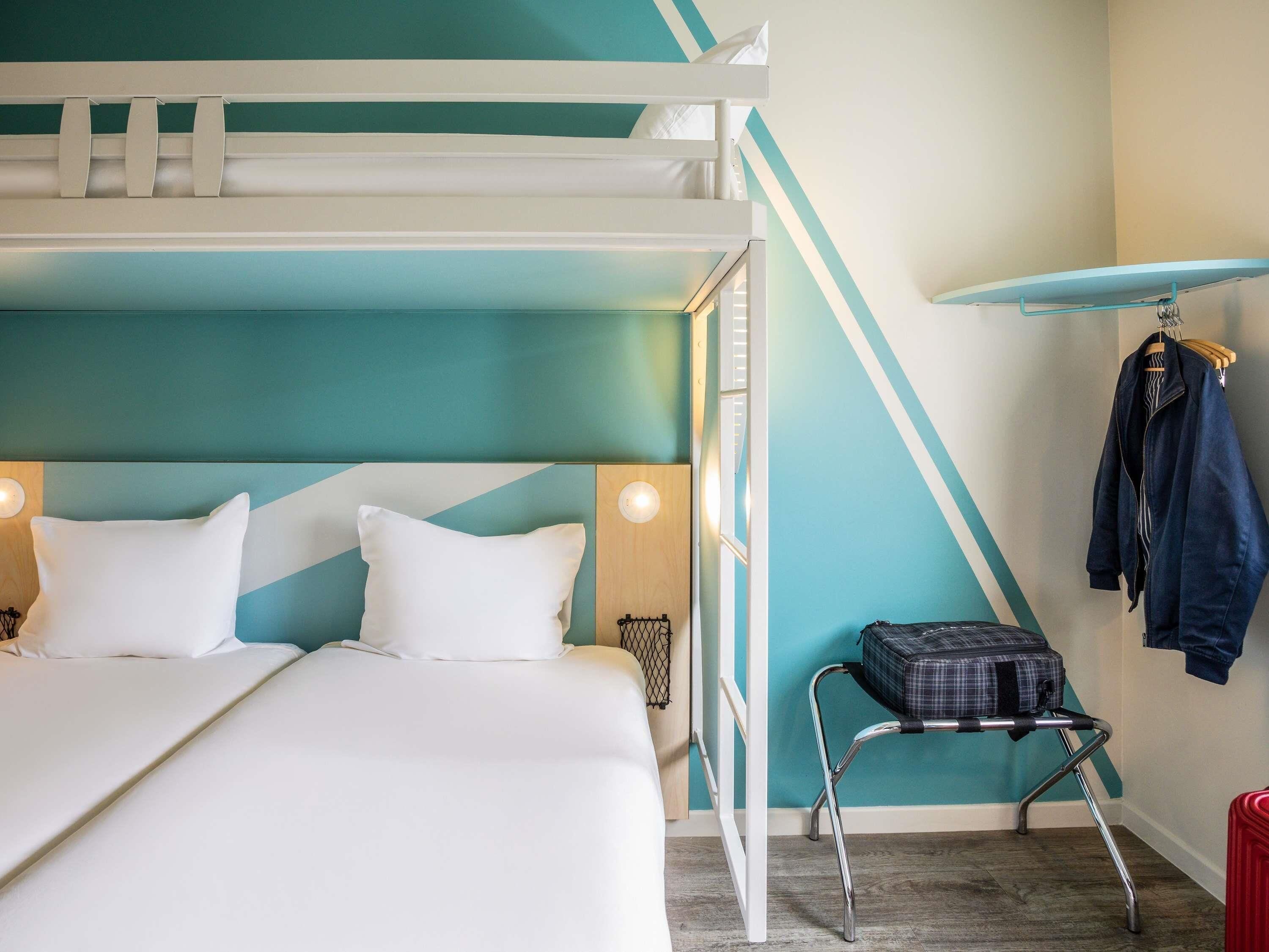 A bunk bed with white bedding, a small table with a checkered bag, and a coat rack with a jacket.